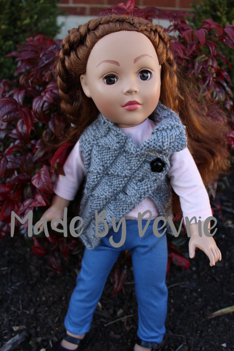 K&ouml;nnte beinhalten: Eine Puppe tr&auml;gt eine graue Strickweste mit einem schwarzen Knopf, ein rosa Langarmshirt, blaue Jeans und eine braune geflochtene Frisur. Die Puppe steht vor einem Hintergrund aus roten Bl&auml;ttern.