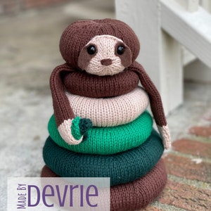 May include: A brown knitted sloth with a pink face and green and dark green rings stacked on top of it. The sloth is sitting on a brick surface. The text "MADE BY Devrie Digital Download" is at the bottom of the image.