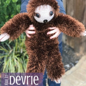 May include: A brown knitted sloth plush toy with a fluffy texture. The sloth is being held by a person wearing blue jeans. The text "MADE BY Devrie Digital Download" is visible in the image.