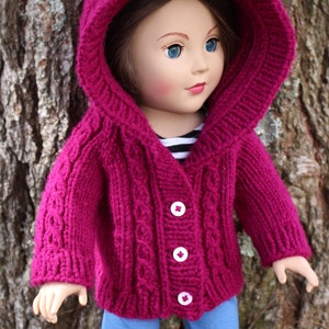 Puede incluir: Una mu&ntilde;eca con una rebeca con capucha de punto magenta con botones blancos y vaqueros azules. La rebeca tiene un dise&ntilde;o de punto trenzado en la parte delantera y en las mangas. La mu&ntilde;eca tiene ojos azules y est&aacute; delante de un tronco de &aacute;rbol.