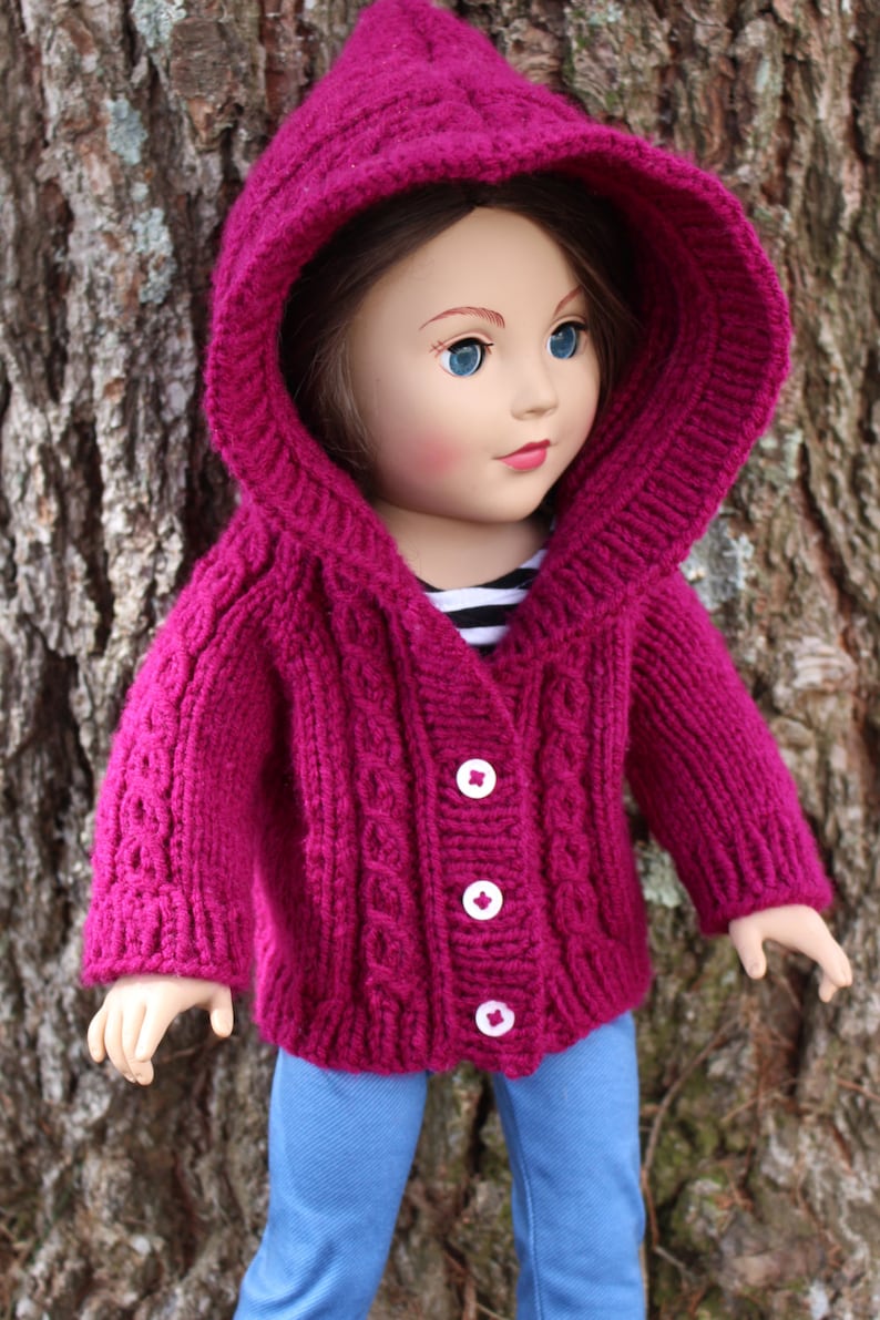 Puede incluir: Una mu&ntilde;eca con una rebeca con capucha de punto magenta con botones blancos y vaqueros azules. La rebeca tiene un dise&ntilde;o de punto trenzado en la parte delantera y en las mangas. La mu&ntilde;eca tiene ojos azules y est&aacute; delante de un tronco de &aacute;rbol.