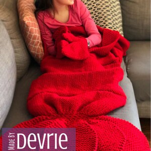 May include: A red knitted mermaid tail blanket with a girl sitting on a couch. The blanket has a large tail fin and a smaller fin on each side. The text "MADE BY Devrie Digital Download" is on the bottom left corner of the image.