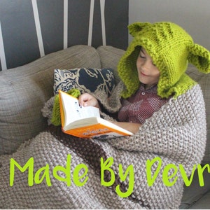 Könnte beinhalten: Ein Kind trägt eine grüne gestrickte Yoda-Mütze und eine braune gestrickte Decke und sitzt auf einem Sofa und liest ein Buch. Der Text "Made By Devrie" ist in der unteren rechten Ecke des Bildes sichtbar.