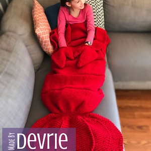 May include: A red knitted mermaid tail blanket with a girl sitting on a couch. The blanket is made with a textured stitch and has a large tail fin. The text "MADE BY Devrie Digital Download" is visible in the bottom left corner of the image.