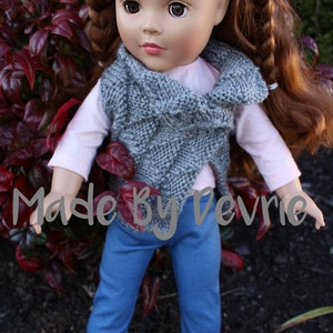 K&ouml;nnte beinhalten: Eine Puppe mit langen braunen Haaren, die auf beiden Seiten geflochten sind, tr&auml;gt eine graue Strickweste, ein rosa Langarmshirt, blaue Jeans und schwarze Schuhe. Die Puppe steht vor einer Pflanze mit roten Bl&auml;ttern. Der Text "Made By Devrie" ist im Bild sichtbar.