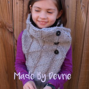 Peut inclure: Une jeune fille portant une chemise violette et un gilet en tricot gris à col roulé avec des boutons. Le gilet a un motif texturé et la fille tient un petit objet brun dans ses mains. Le texte "Made By Devrie" est visible sur l'image.