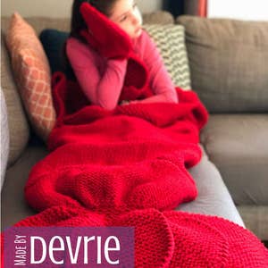 May include: A red knitted blanket with a mermaid tail design. The blanket is on a couch with a young girl sitting on it. The text "MADE BY Devrie Digital Download" is on the blanket.
