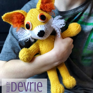 May include: A yellow knitted fox toy with orange ears and a white beard. The fox has large black eyes with white buttons. The toy is being held by a person wearing a gray shirt and blue jeans. The text "MADE BY Devrie Digital Download" is visible at the bottom of the image.