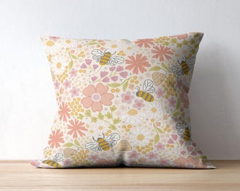 May include: A cream-colored throw pillow with a floral pattern featuring pink, yellow, and white flowers and bees. The pillow has a subtle embroidered design.