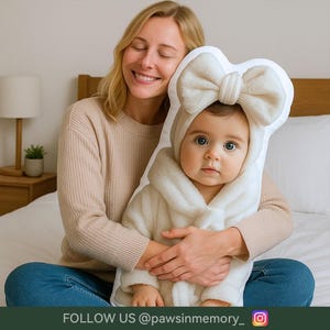 May include: A plush, cream-colored pillow shaped like a baby in a hooded robe and bow. The pillow features a photo of a baby with blue eyes. The text "FOLLOW US @pawsinmemory_" is at the bottom.
