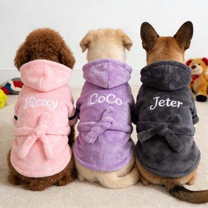 May include: Three dogs wearing personalized hooded robes. The robes are in pink, purple, and gray, each with a matching belt and the names "Roxy", "CoCo", and "Jeter" embroidered in white. The dogs are facing away from the camera.
