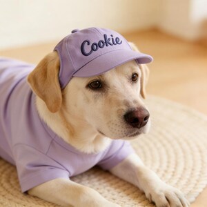 May include: A light-colored Labrador Retriever wearing a matching lavender baseball cap and t-shirt. The cap has the name "Cookie" embroidered in a dark blue script. The dog is lying on a woven mat.