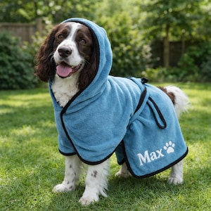 May include: A dog wearing a light blue hooded robe with black trim, personalized with the name "Max" and a paw print. The dog has brown and white fur and is standing on green grass.