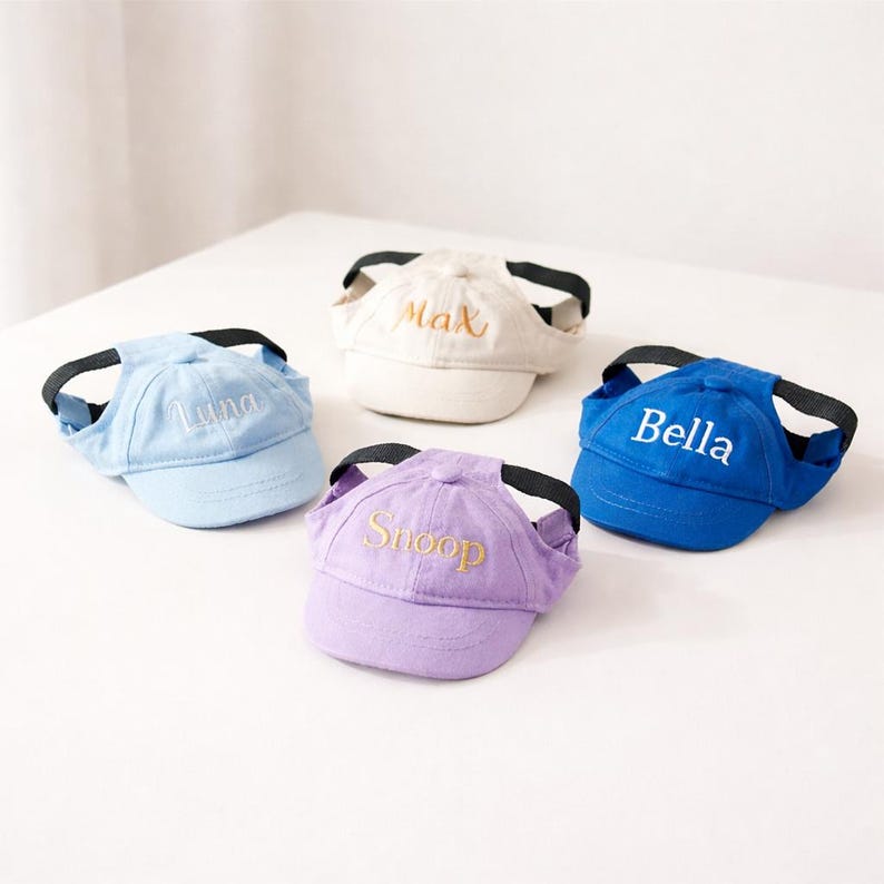 May include: Four pet baseball caps in pastel colors: light blue, cream, purple, and royal blue. Each cap has a black strap and embroidered name: "Luna", "Max", "Snoop", and "Bella" in matching thread. The caps are designed for small dogs or cats.