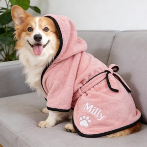May include: A smiling corgi dog wearing a pink hooded bathrobe with black trim. The bathrobe has a matching belt and the name "Milly" embroidered on the front, along with a paw print design.