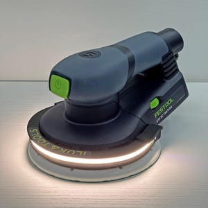 Op de afbeelding: Een grijze en zwarte elektrische schuurmachine met een groene aan/uit-knop. De schuurmachine heeft de tekst "FESTOOL ETS EC 150/5 EQ" op de zijkant staan. De schuurzool is wit en heeft de tekst "ILUKA TOOLS" erop.