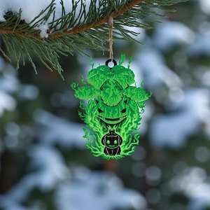 May include: A green and black ornament featuring a stylized creature design. The ornament is hanging from a natural twine string against a snowy background. The design has a glowing green outline and a black interior.