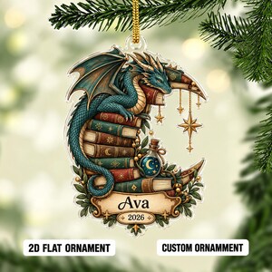 May include: A flat acrylic ornament featuring a teal dragon perched on a stack of colorful books shaped like a crescent moon. The design includes hanging gold stars, a small potion bottle, and a banner with the name Ava and the year 2026.