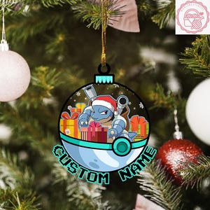 May include: A round Christmas ornament featuring a blue cartoon character wearing a Santa hat, surrounded by wrapped gifts. The ornament has the words "CUSTOM NAME" in a stylized font. The ornament is hanging on a Christmas tree.