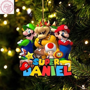 May include: A vibrant Christmas decoration featuring characters from the Super Mario video game series. The ornament includes Mario, Luigi, Bowser, Toad, and the text "SUPER DANIEL". It hangs from a gold string.
