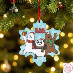 May include: A snowflake-shaped Christmas ornament featuring the We Bare Bears characters. The ornament shows a polar bear, a panda, and a brown bear, all wearing Santa hats and scarves. The ornament is light blue with snowflakes.