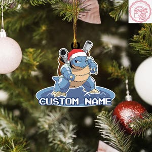 May include: A blue and tan cartoon character ornament wearing a Santa hat. The ornament has the text "CUSTOM NAME" in white pixelated letters. The ornament is hanging on a Christmas tree with other ornaments.