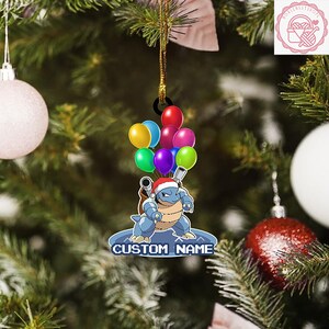 May include: A Christmas tree ornament featuring a blue cartoon character with a Santa hat, holding colorful balloons. The ornament has the text "CUSTOM NAME" at the bottom. The background includes a Christmas tree with ornaments.