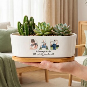 Könnte beinhalten: Ein weißer, ovaler Keramikübertopf mit Bambusuntersetzer, bepflanzt mit einem Kaktus und zwei Sukkulenten. Der Übertopf zeigt das Wort DAD mit Fotos einer Person und eines Kindes in den Buchstaben sowie den Text To the world you are a dad, but to our family you are the world.