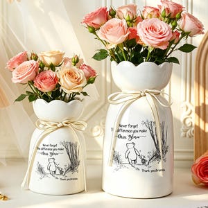 May include: Two white ceramic vases with scalloped rims, each tied with a beige ribbon. The vases feature a black line drawing of a bear and a piglet walking, with the text: Never forget the difference you make, Mrs. Ylnw, Thank you Brenda.