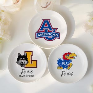 May include: Three white ceramic plates with college logos. One plate has a red and blue "A" logo with "AMERICAN" text. Another features a wolf and "L" with "Rachel CLASS OF 2025". The third plate has a blue bird logo with "KU" and "Rachel".