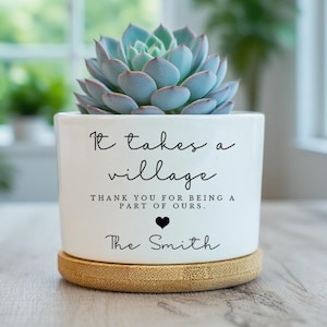 May include: A white ceramic plant pot with a succulent. The pot is inscribed with "It takes a village" and "Thank you for being a part of ours." with a heart symbol and "The Smith" below. It rests on a bamboo coaster.