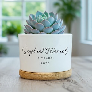 May include: A white ceramic planter with a succulent plant. The planter has the names "Sophia & Daniel", "8 YEARS", and "2025" in black script. The planter sits on a round, natural-colored bamboo coaster. The succulent has blue-green leaves.