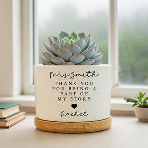 Puede incluir: Una maceta de cerámica blanca con una planta suculenta. La maceta tiene el texto "Sra. Smith, Gracias por ser parte de mi historia, Rachel" en escritura negra. La maceta se asienta sobre un posavasos redondo de bambú marrón claro.