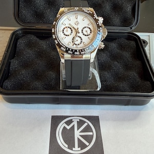 May include: A silver-toned wristwatch with a white face, black subdials, and a black rubber strap. The watch is displayed in a black case with foam padding. The watch face has the text "Units Per Hour". The logo "XIXI Time Pieces" is visible.