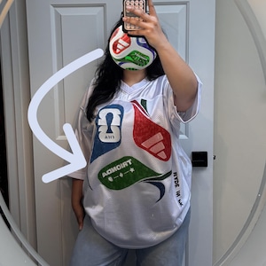 May include: A white mesh jersey featuring a V-neckline and short sleeves. The front displays large, colorful graphic patches in red, blue, and green with the text FIFA and TRIONDA. The side of the jersey includes the text MADE IN PK.