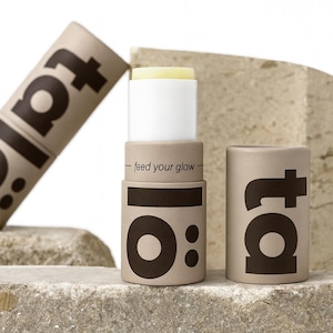 May include: A skincare product with a white and yellow balm is displayed. The product is in a tan cylindrical container with the text "ta" and "10:" in dark brown. The words "feed your glow" are also printed on the container.