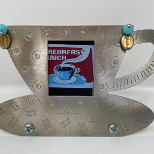 May include: A silver metal picture frame shaped like a coffee cup and saucer. The frame has a small picture inside of a vintage coffee advertisement that says "Breakfast Lunch". The frame is decorated with small blue and gold accents.