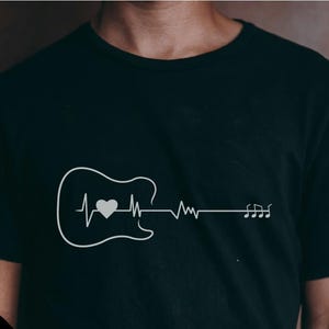 Puede incluir: Camiseta negra con un gráfico blanco de una guitarra, una línea de latidos y notas musicales. El diseño incluye un corazón dentro del contorno de la guitarra. La esquina inferior izquierda tiene el texto "ART INVADER" y opciones de tipo de archivo.