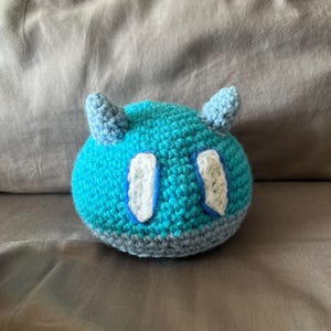 Puede incluir: Un juguete de peluche hecho a mano, tejido a ganchillo, con forma de criatura redonda. El juguete es principalmente turquesa con cuernos azul claro y detalles blancos y azules. La base es gris.