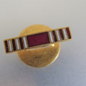 Könnte beinhalten: Eine goldfarbene Anstecknadel mit einem rot-weiß gestreiften Banddesign. Die Nadel ist rund und hat einen erhöhten Rand.