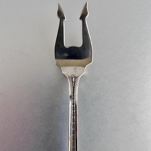 May include: A silver-toned metal serving fork featuring two distinct, pointed tines with a curved, flared design. The handle displays a vertical, grooved pattern with an ornate, embossed decorative element at the base of the fork head.