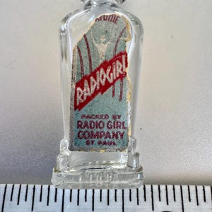 May include: A small, clear glass perfume bottle with a black cap. The label reads "Radio Girl" in red lettering over a blue background. The bottle is marked "Packed by Radio Girl Company, St. Paul."