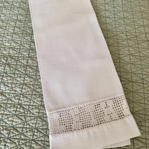 Puede incluir: Toalla de mano blanca con tejido texturizado y borde calado decorativo. La toalla doblada descansa sobre una superficie acolchada verde. Fabricada con un material suave y absorbente.