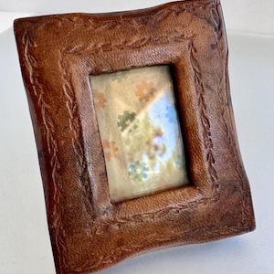 May include: A rectangular brown leather picture frame with a textured surface and a decorative embossed vine pattern along the inner border. The frame contains a light-colored insert featuring a repeating pattern of small orange, green, and blue flowers.