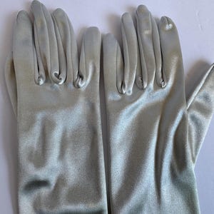 May include: A pair of light gray satin gloves. The gloves are smooth and shiny, with a slight sheen. The fingers are neatly tailored, and the gloves appear to be unworn. The gloves are likely for formal wear or a costume.