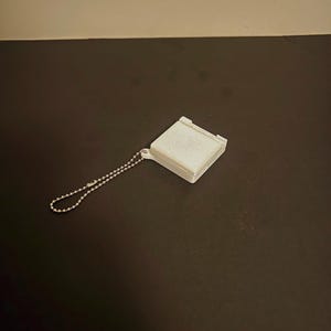 May include: A small, white, square-shaped object with a textured surface and a silver ball chain attached. The object appears to be a small container or case, possibly for holding small items. The background is a dark surface.