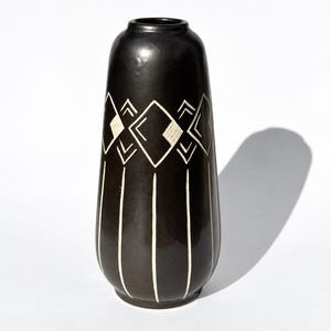 Pode incluir: Um vaso cilíndrico alto de cerâmica com um esmalte liso castanho escuro, quase preto. A secção superior apresenta um padrão repetitivo de formas geométricas de diamante e linhas brancas, enquanto a inferior tem linhas verticais brancas.