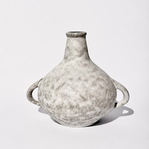 Könnte beinhalten: Eine strukturierte, graue Keramikvase mit bauchigem Körper und zwei Schlaufengriffen. Die Vase hat einen schmalen Hals und einen dunklen Rand. Die Oberfläche hat ein gesprenkeltes Aussehen und die Vase steht auf weißem Hintergrund.
