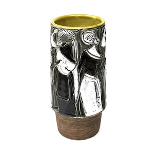 May include: A cylindrical ceramic vase featuring a relief design of stylized figures in black and white against a dark gray background. The interior rim is finished in a metallic gold glaze, while the base has a textured, brown, wood-like finish.