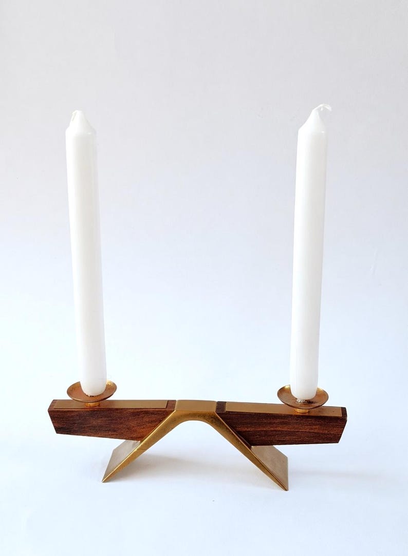 K&ouml;nnte beinhalten: Ein Kerzenhalter im Mid-Century-Modern-Stil mit zwei wei&szlig;en Kerzen. Der Halter hat einen dunklen Holzsockel mit Messingakzenten und zwei Messing-Kerzenschalen. Das Design ist minimalistisch und elegant, passend f&uuml;r die Inneneinrichtung.